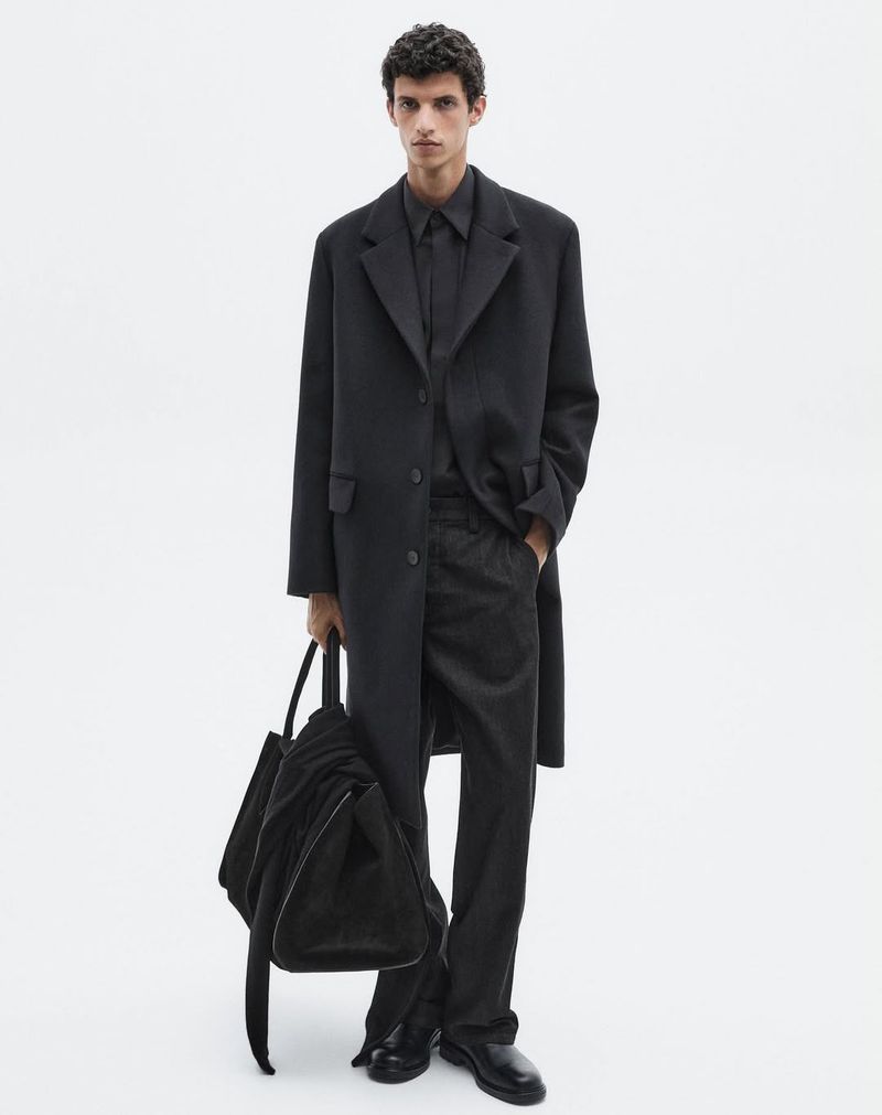 Fashion model standing in an all-black tailored coat, trousers, and shirt while holding a large black weekender bag against a white seamless background.
