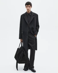 Fashion model standing in an all-black tailored coat, trousers, and shirt while holding a large black weekender bag against a white seamless background.