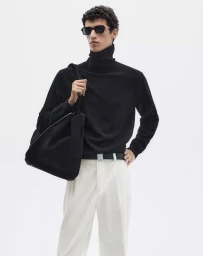 Male model wearing a black turtleneck sweater, white trousers, and dark sunglasses carrying a black shoulder tote against a white backdrop.