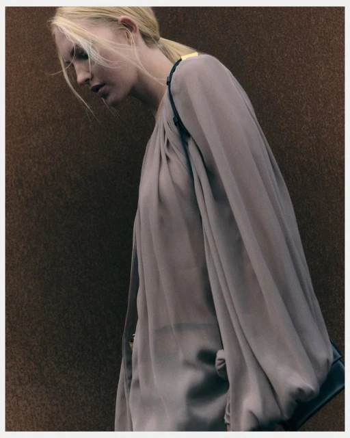 Blonde woman in a draped, long-sleeved beige dress against a textured brown background in a studio-style fashion portrait.