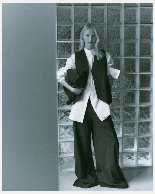 A fashion shot featuring a fair-skinned adult female model in a white shirt, vest, and wide-leg black pants, holding a hat against a glass block wall.