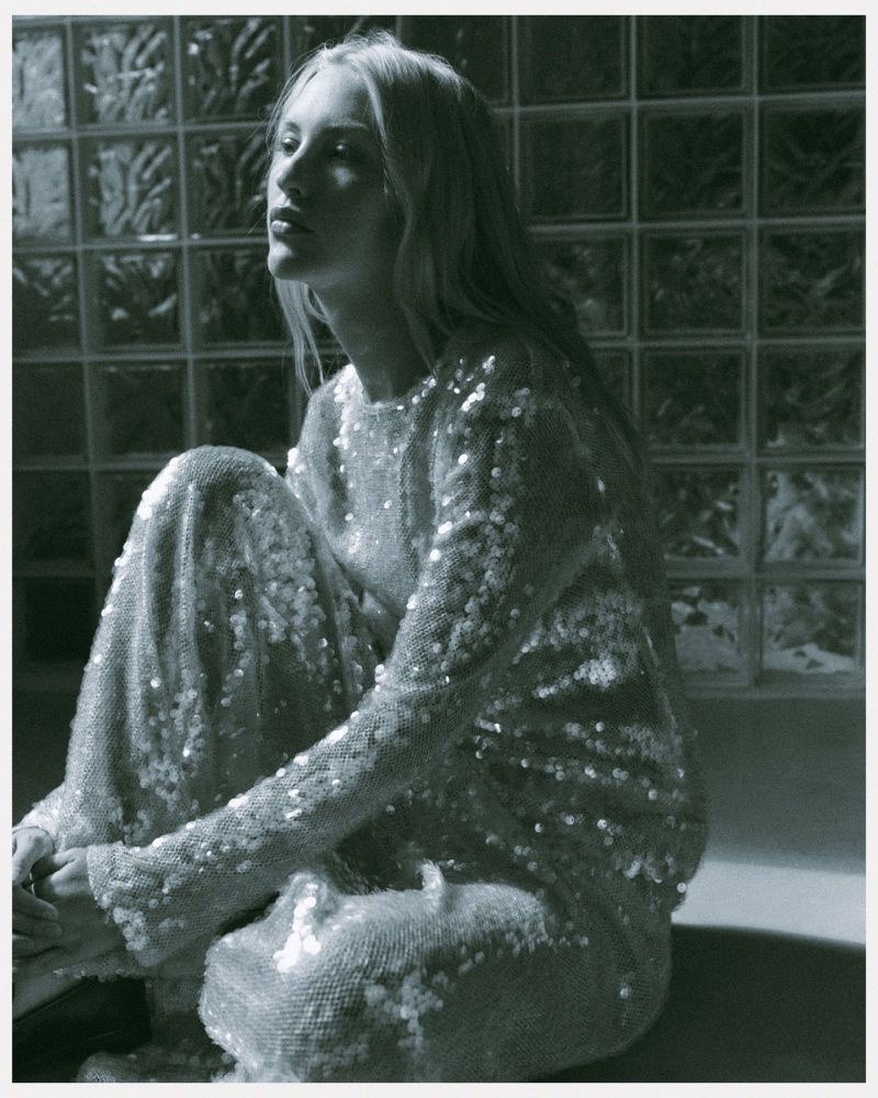 Monochromatic fashion portrait of a poised, slender blonde woman in a shimmering sequin dress, seated against a textured glass block wall.