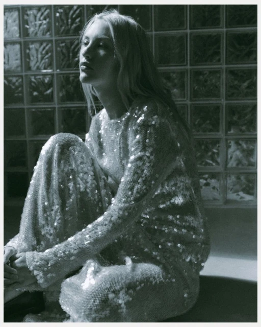 Monochromatic fashion portrait of a poised, slender blonde woman in a shimmering sequin dress, seated against a textured glass block wall.