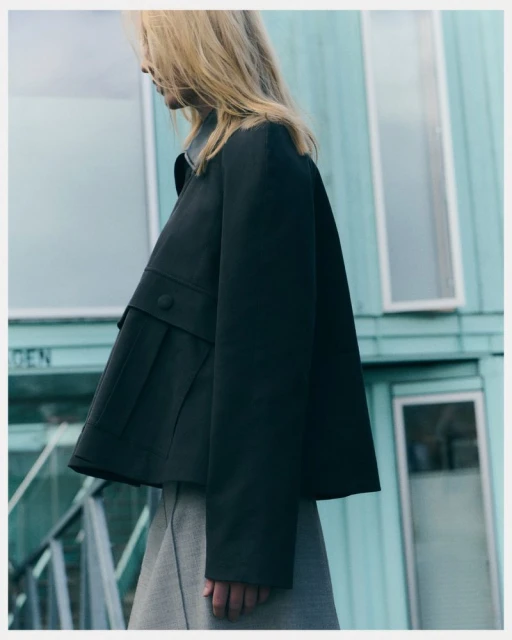 Fashionable woman in a dark coat and light skirt stands outside a light blue building with white-framed windows in an eye-level shot.