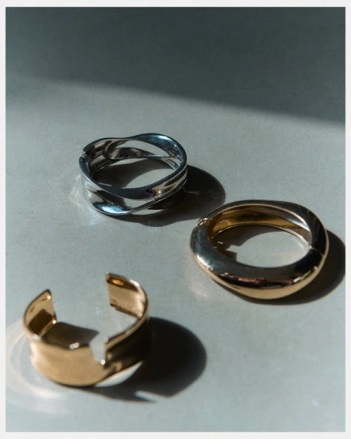 Three metal bracelets—two gold and one silver—are arranged on a neutral surface with contrasting shadows, showcasing modern jewelry design.