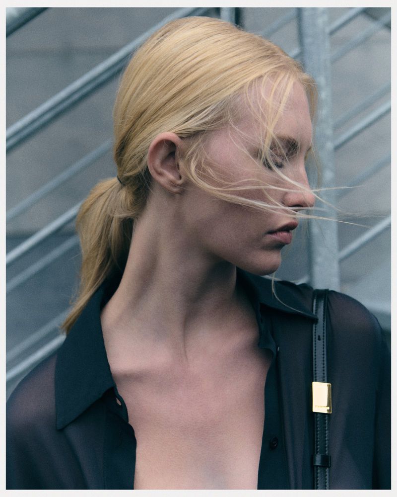 Close-up portrait of a blonde woman in a sheer black blouse against a grey industrial background with soft, diffused lighting.