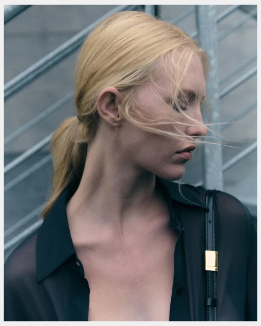 Close-up portrait of a blonde woman in a sheer black blouse against a grey industrial background with soft, diffused lighting.