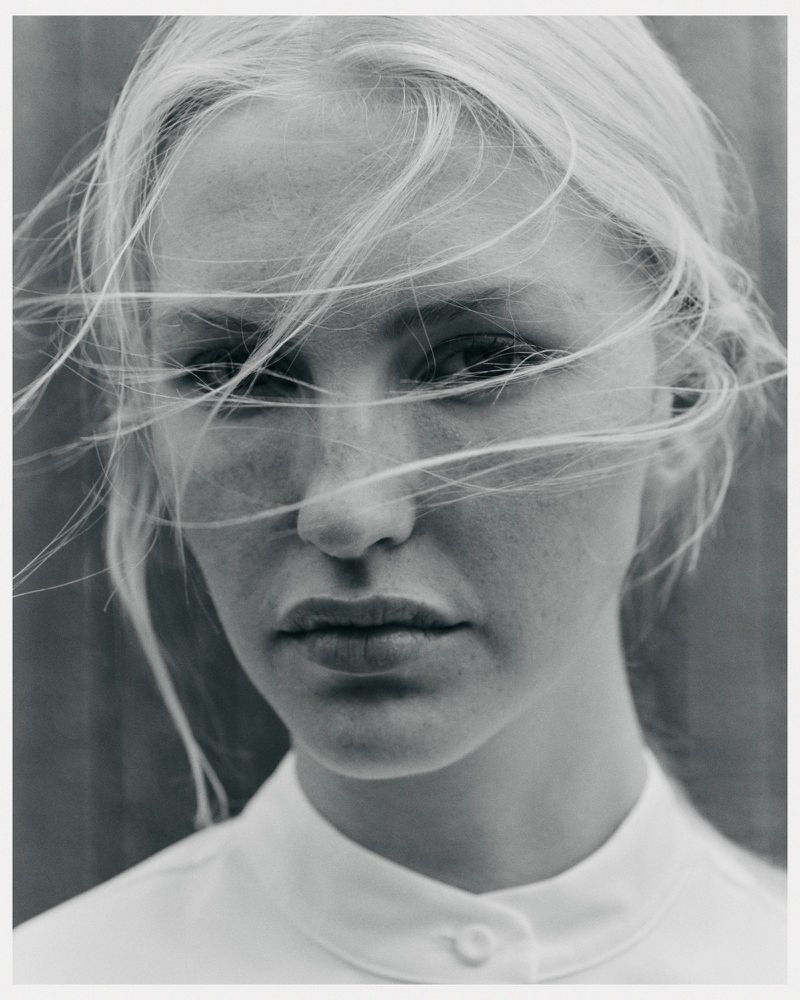 Striking black and white portrait of a blond woman with windblown hair, wearing a collared shirt, set against a neutral background.