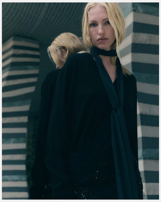 Model with blonde hair wearing a black top and scarf standing against striped architectural pillars