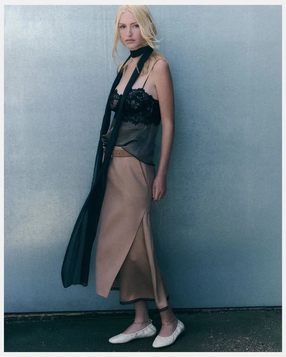 Model Molly Merland posing in a black lace camisole top, long skinny scarf, beige midi skirt, and white ballet flats against a muted wall.