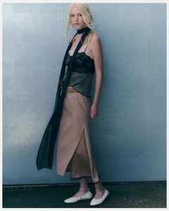 Model Molly Merland posing in a black lace camisole top, long skinny scarf, beige midi skirt, and white ballet flats against a muted wall.