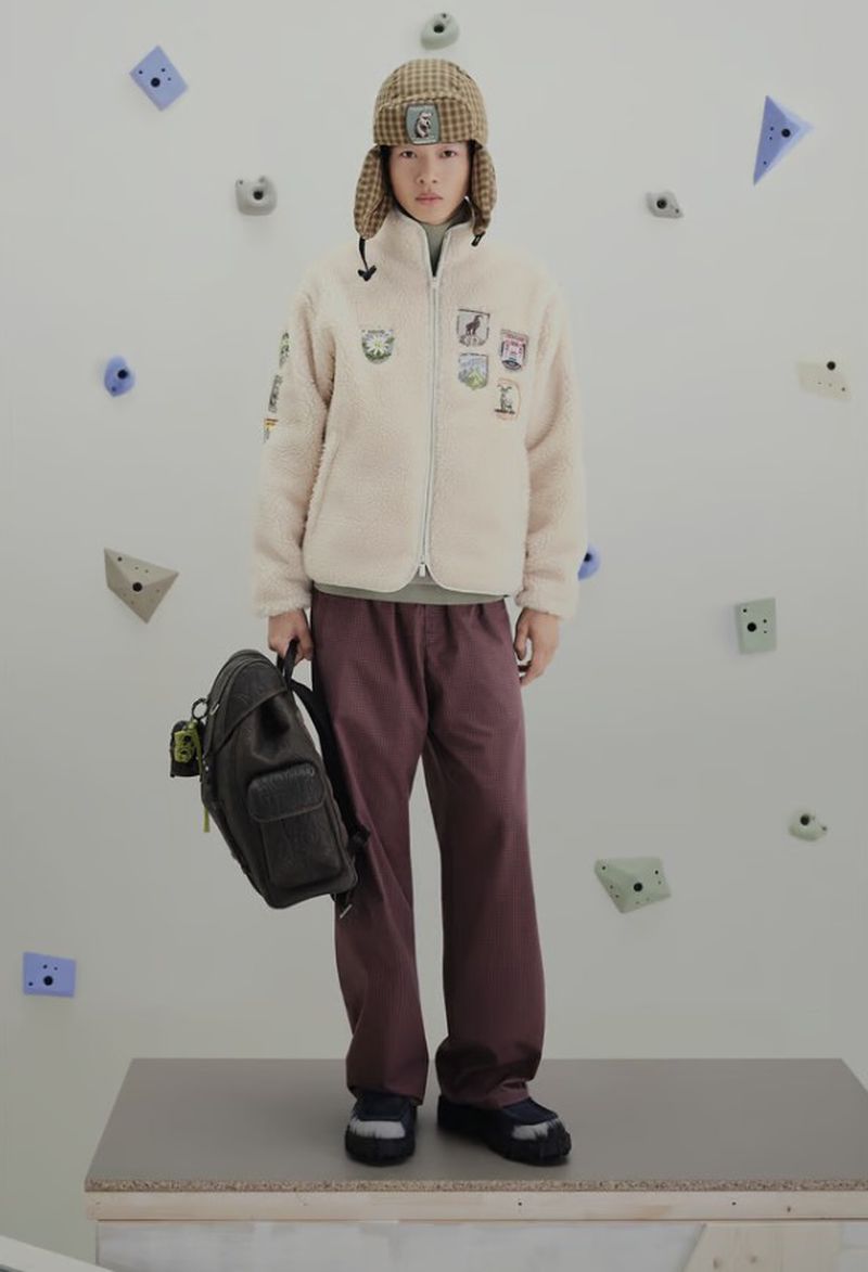 Full shot of a young adult in streetwear, posing in a cream sherpa jacket, plum pants, and a patterned hat, holding a backpack.