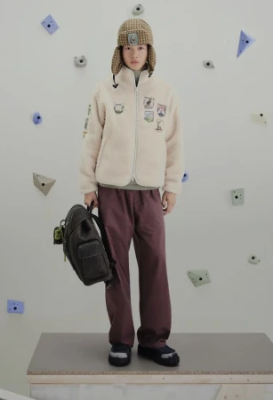 Full shot of a young adult in streetwear, posing in a cream sherpa jacket, plum pants, and a patterned hat, holding a backpack.