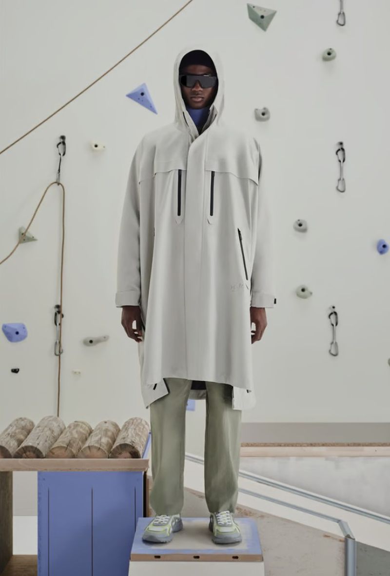 A tall male fashion model in a hooded overcoat and sunglasses stands on a platform in front of a climbing wall backdrop.