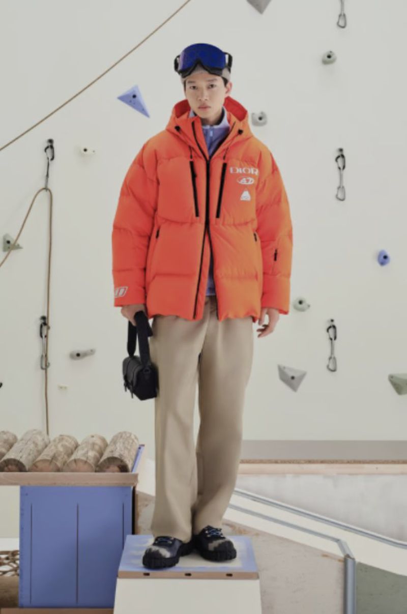 A young adult stands on a platform wearing an orange puffer jacket, beige pants, and ski goggles in front of a climbing wall.