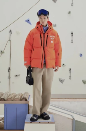 A young adult stands on a platform wearing an orange puffer jacket, beige pants, and ski goggles in front of a climbing wall.