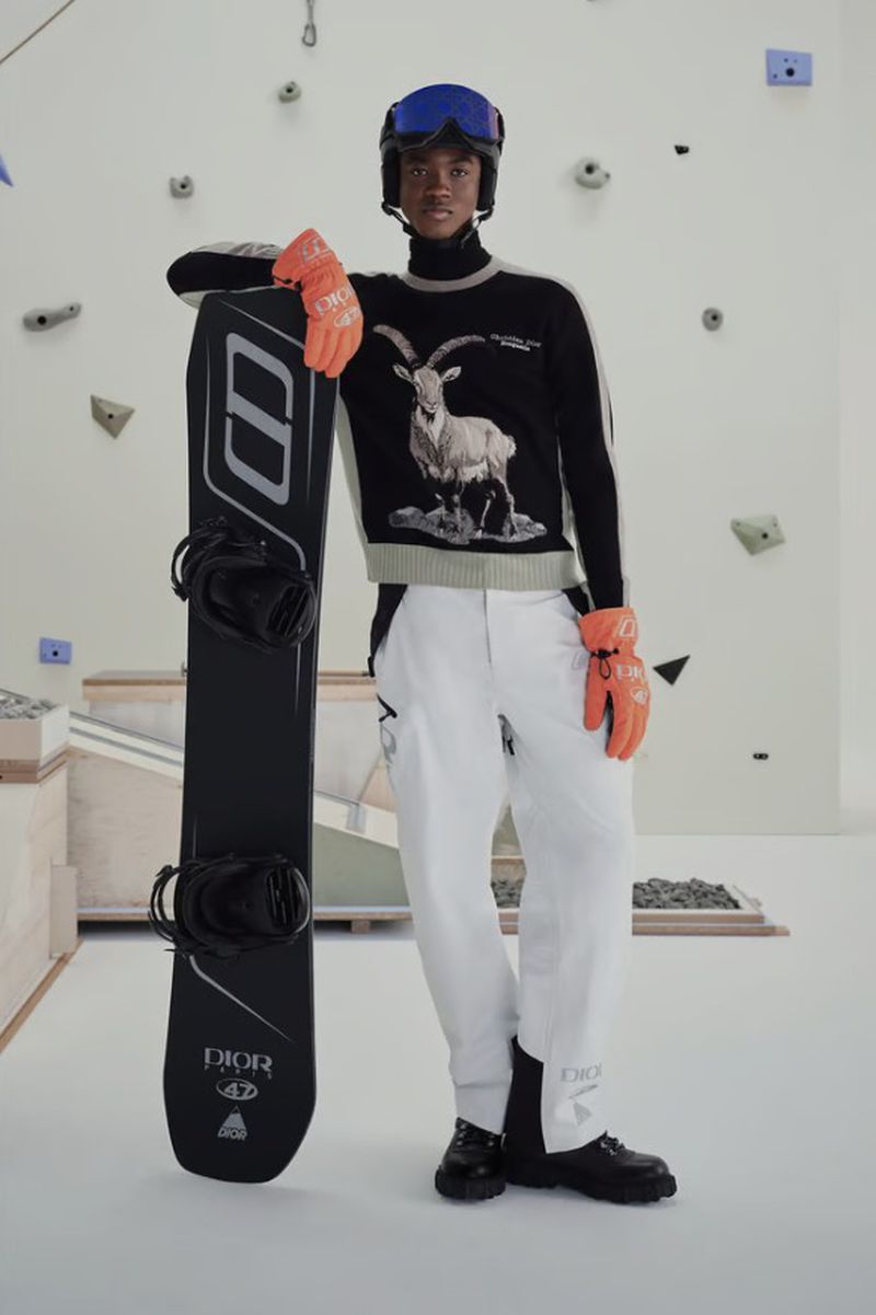 Man in winter fashion wearing ski goggles, sweater with goat graphic, and white pants, leaning on a snowboard, indoor setting.