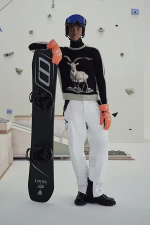 Man in winter fashion wearing ski goggles, sweater with goat graphic, and white pants, leaning on a snowboard, indoor setting.