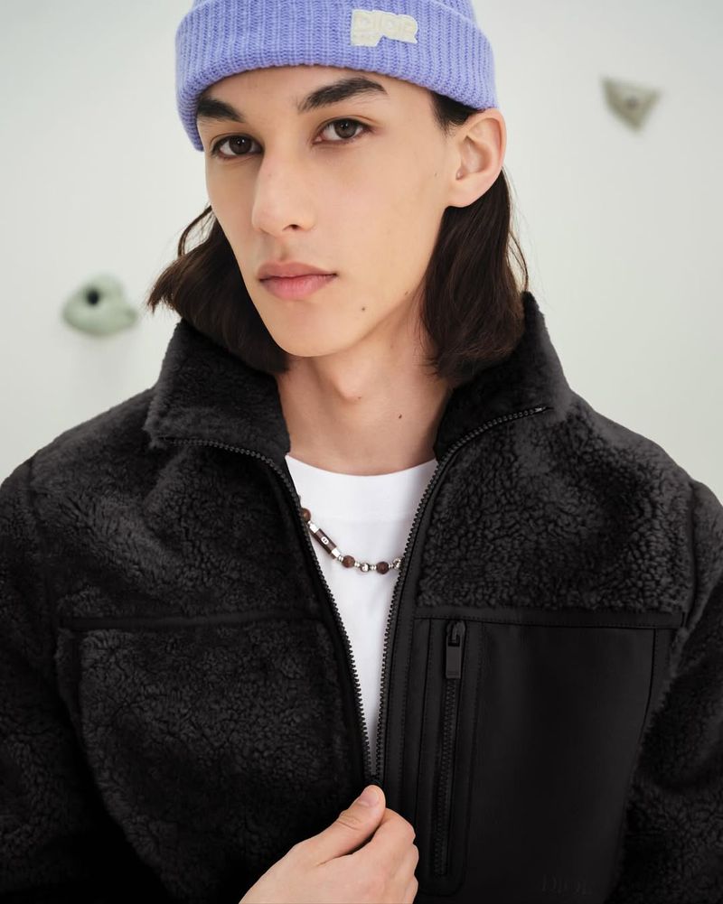 Young adult male wearing a lavender beanie and black sherpa jacket posing indoors against a white wall with climbing holds.