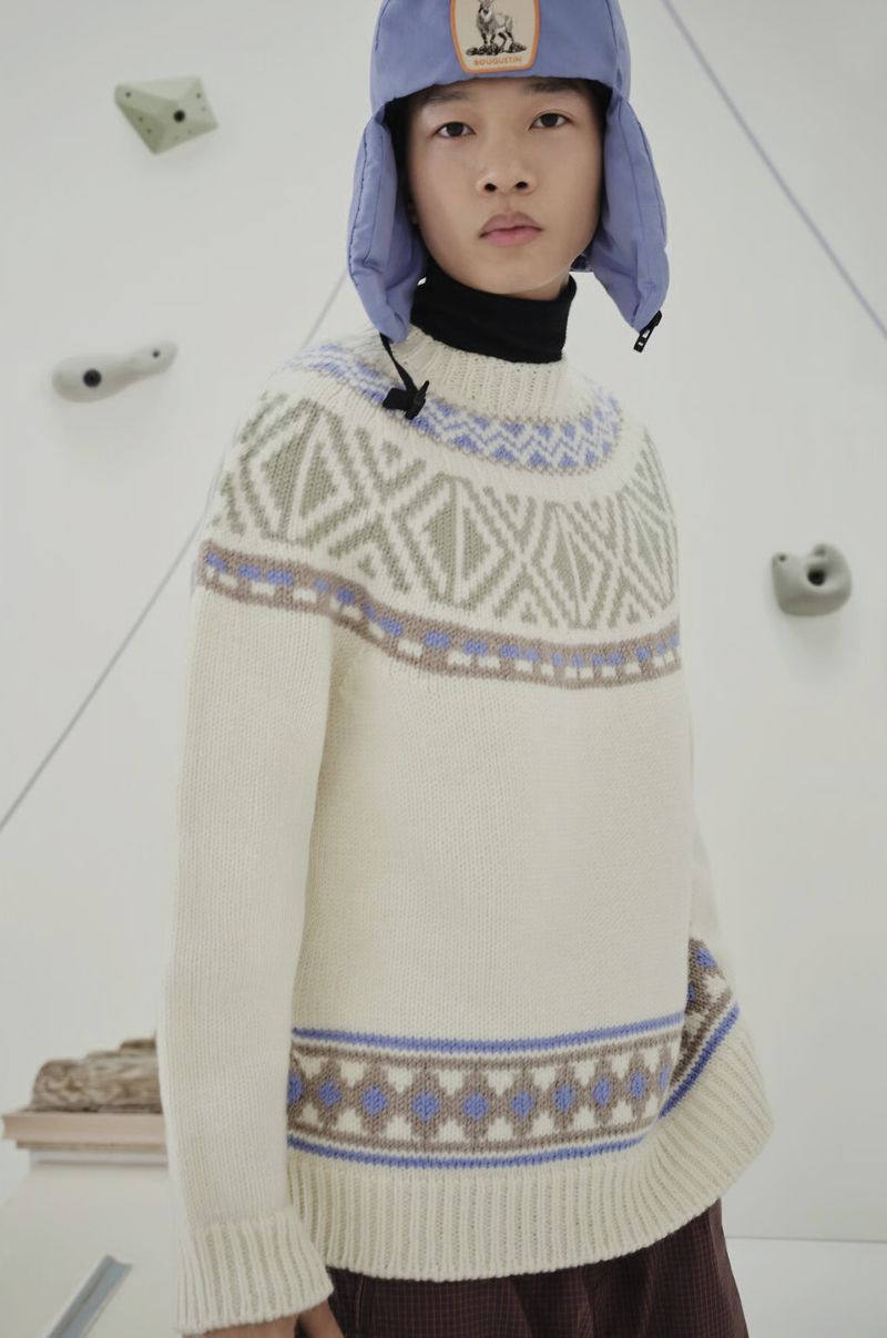 Young adult wears a patterned sweater and earflap hat against a climbing wall backdrop in a studio setting, showcasing fashion attire.