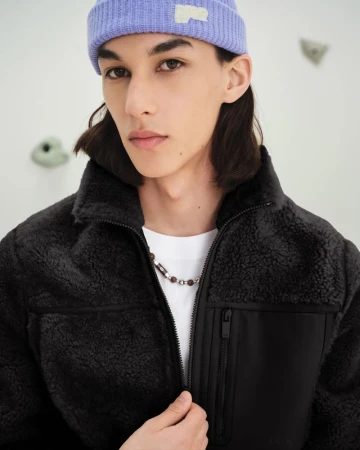 Young man wearing a lilac knit beanie and black textured fleece zip jacket in front of an indoor climbing wall.