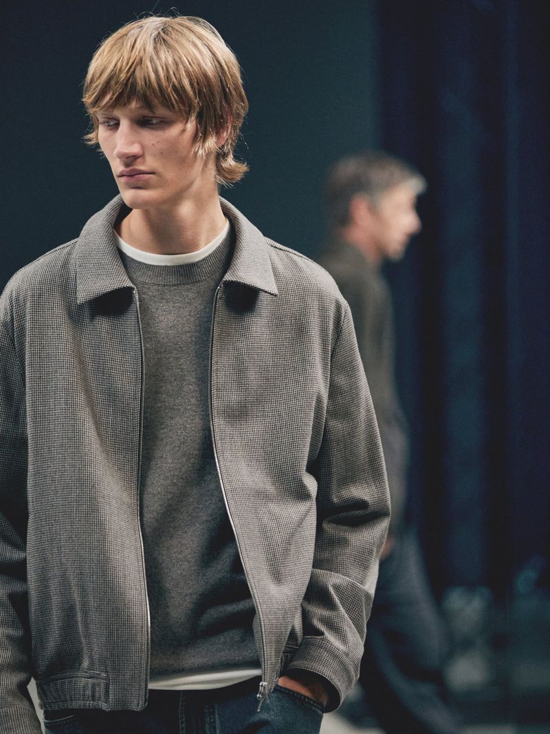 A fair-skinned male model with blond hair poses indoors in a gray jacket and sweater with jeans against a blurred background.