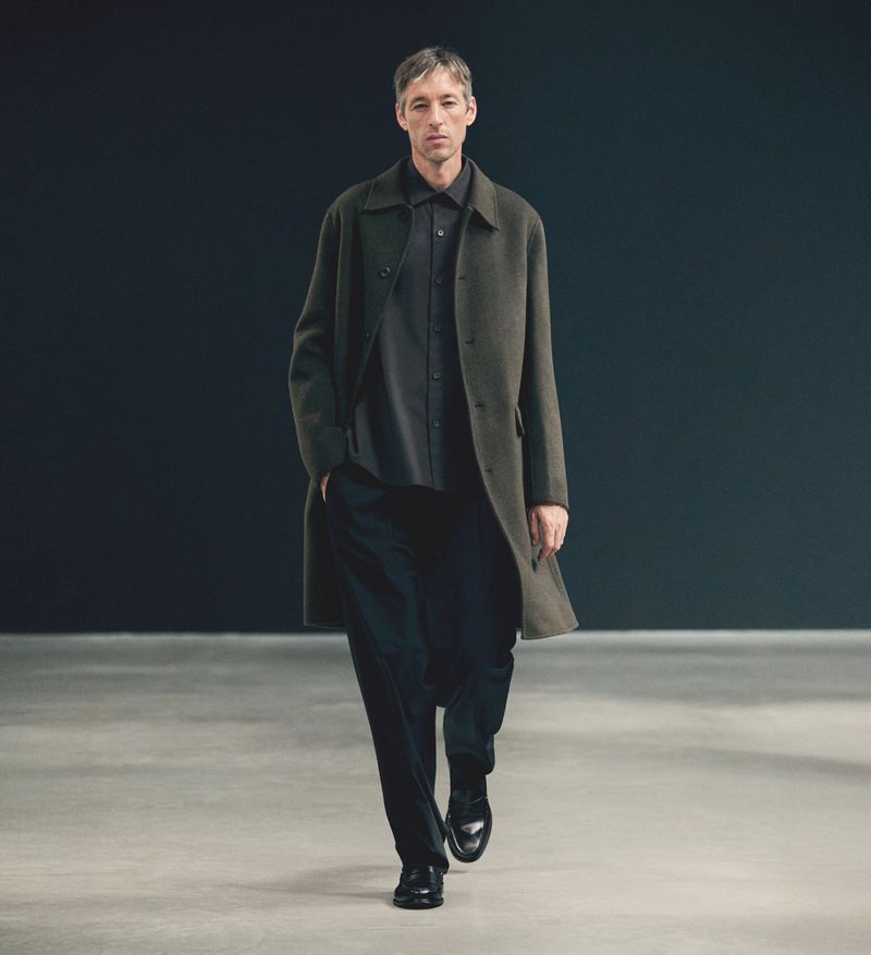 A man wearing a long coat walks forward, presenting a minimalist fashion style in an indoor setting with a neutral backdrop.