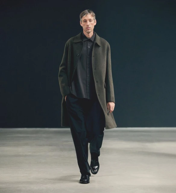 A man wearing a long coat walks forward, presenting a minimalist fashion style in an indoor setting with a neutral backdrop.