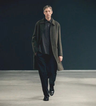 A man wearing a long coat walks forward, presenting a minimalist fashion style in an indoor setting with a neutral backdrop.