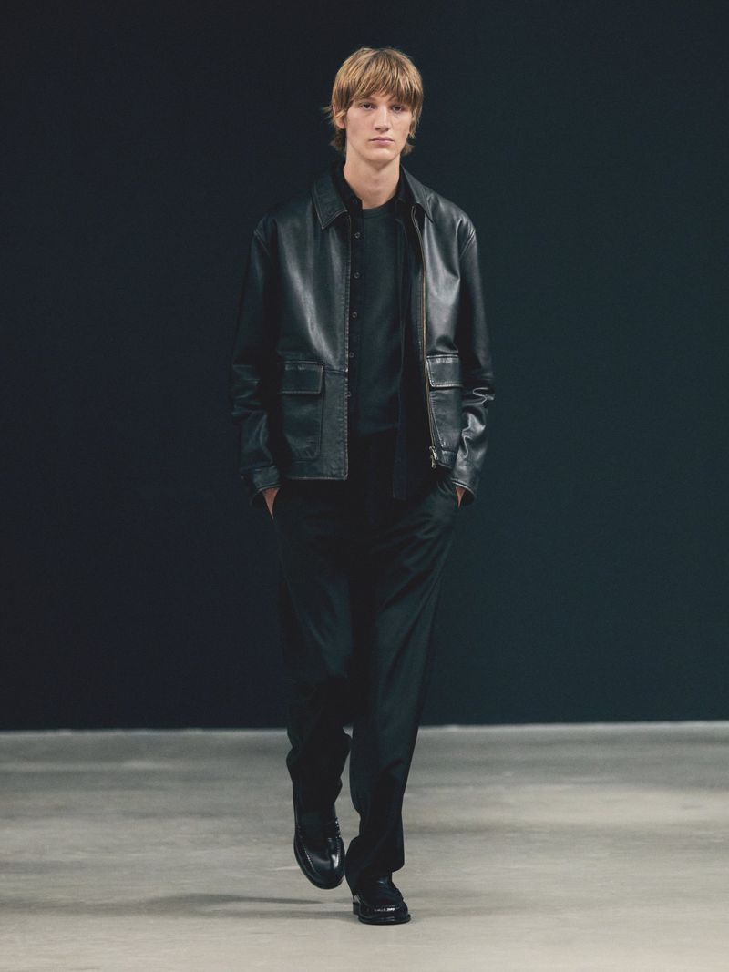 A male fashion model walks toward the camera wearing a black leather jacket, black pants, and black shoes in a dimly lit indoor space.