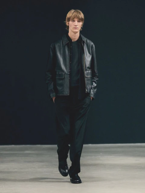 A male fashion model walks toward the camera wearing a black leather jacket, black pants, and black shoes in a dimly lit indoor space.