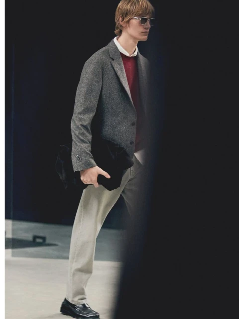 Man in grey blazer, red sweater, and light-wash jeans poses against a dark backdrop, holding a black jacket and wearing sunglasses.