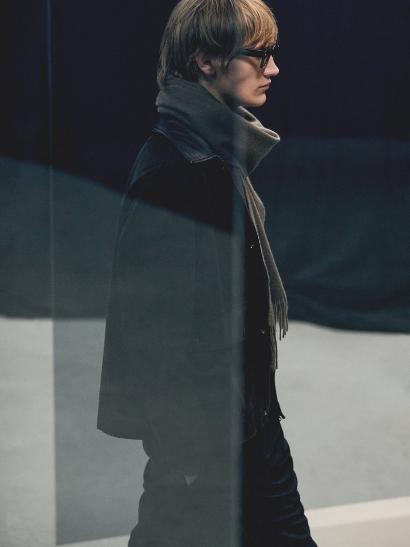 A slender man with blond hair and glasses, wearing a dark coat and grey scarf, poses in a fashion shot with soft lighting.