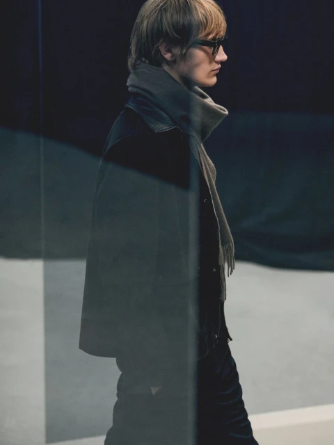 A slender man with blond hair and glasses, wearing a dark coat and grey scarf, poses in a fashion shot with soft lighting.