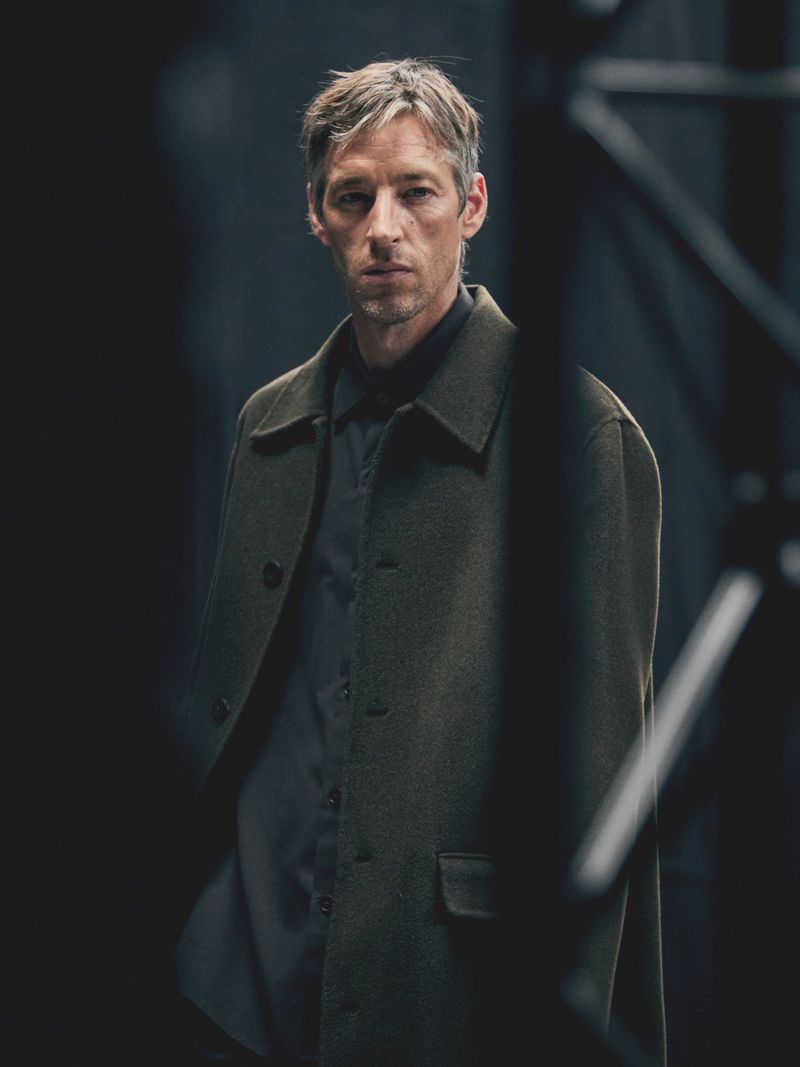 Portrait of an adult male with fair skin and short gray hair, wearing a dark shirt and a green coat, standing indoors in a dark environment.