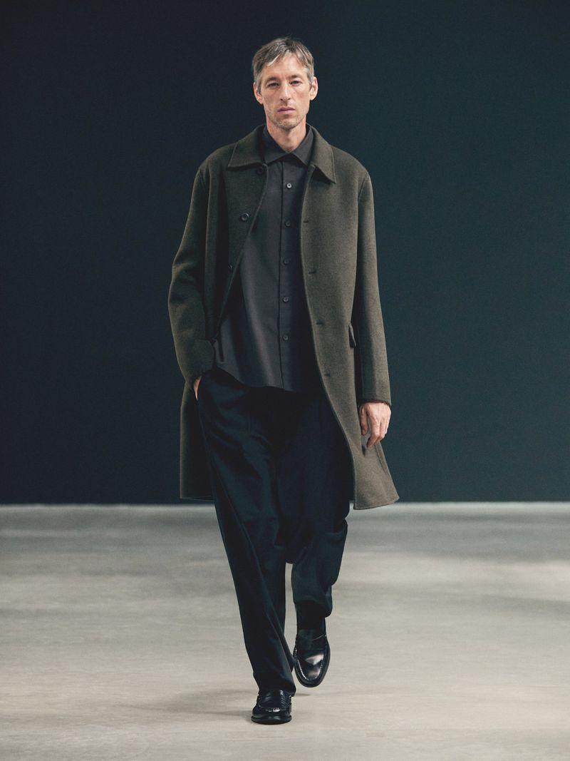 A mature man in a dark shirt and pants, layered with an olive overcoat, walks on a concrete floor against a dark grey backdrop in a studio.