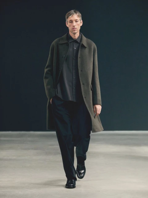 A mature man in a dark shirt and pants, layered with an olive overcoat, walks on a concrete floor against a dark grey backdrop in a studio.