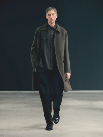 A mature man in a dark shirt and pants, layered with an olive overcoat, walks on a concrete floor against a dark grey backdrop in a studio.