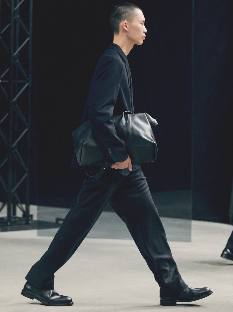 A male fashion model walks the runway in a dark suit and black leather bag during a fashion show on a gray runway with dark backdrop.