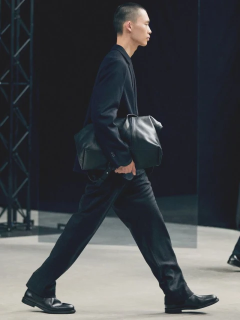 A male fashion model walks the runway in a dark suit and black leather bag during a fashion show on a gray runway with dark backdrop.