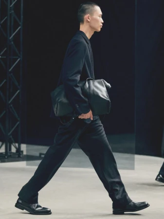 A male fashion model walks the runway in a dark suit and black leather bag during a fashion show on a gray runway with dark backdrop.