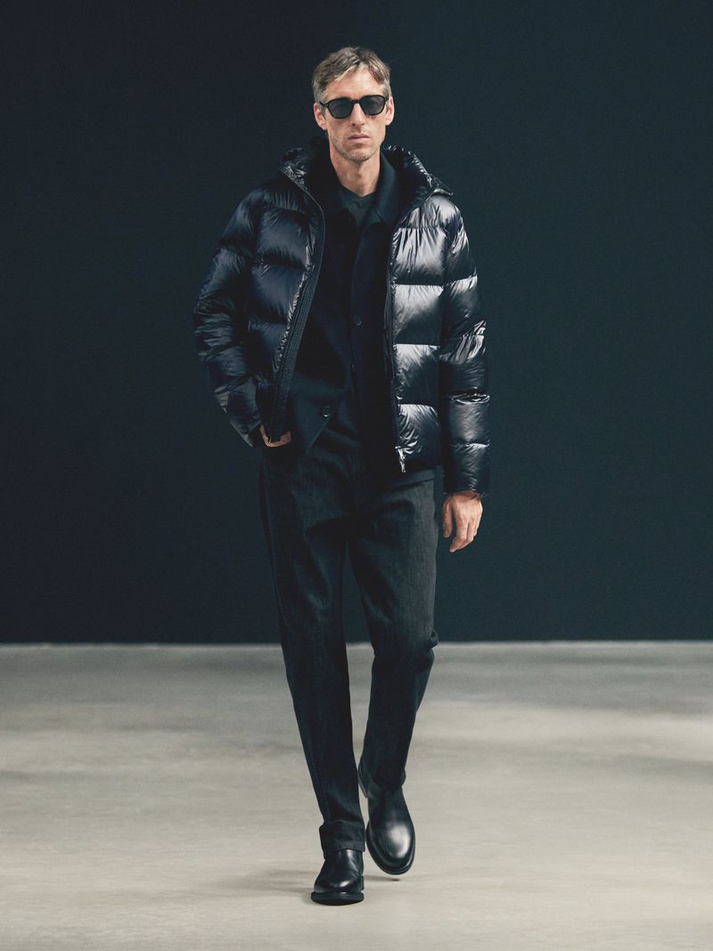 A male model walks wearing a glossy black puffer jacket, dark trousers, black leather boots, and dark sunglasses against a dark backdrop.