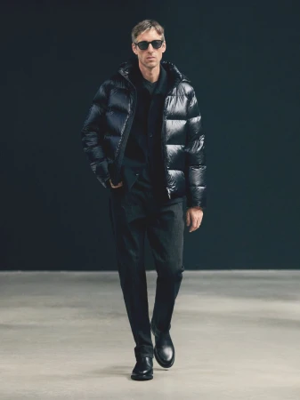 A male model walks wearing a glossy black puffer jacket, dark trousers, black leather boots, and dark sunglasses against a dark backdrop.