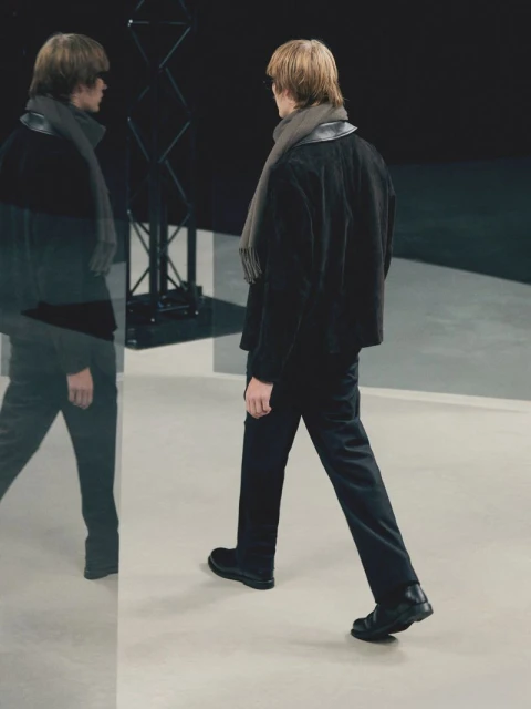 A slender adult male walks in black suit and scarf in a photo studio with his reflection visible in a glass pane.