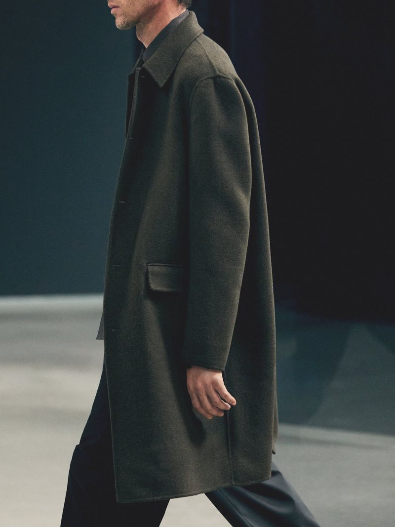 A man walks in a dark overcoat. The fashion shot features a side profile view of the person in shadow with studio lighting.