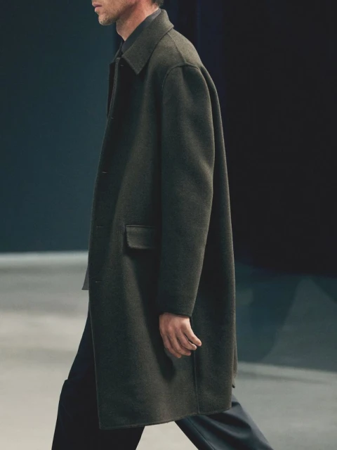 A man walks in a dark overcoat. The fashion shot features a side profile view of the person in shadow with studio lighting.