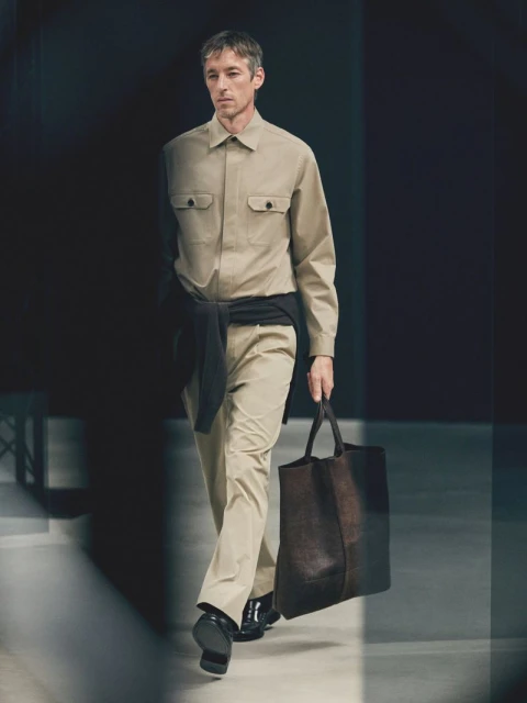 An adult male model walks a runway in a tan collared shirt and pants, carrying a dark tote bag, under studio lighting.