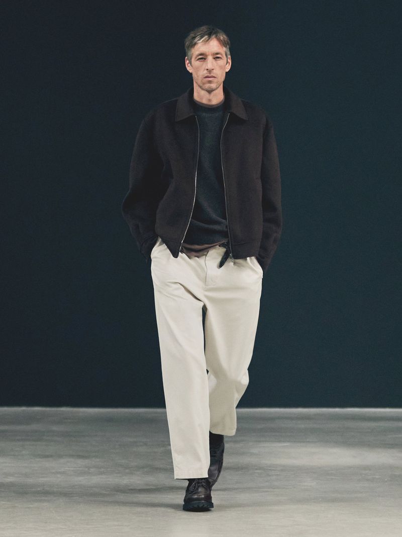 Adult man walks forward in a studio with a dark background wearing a black jacket, sweater, and light-colored pants.