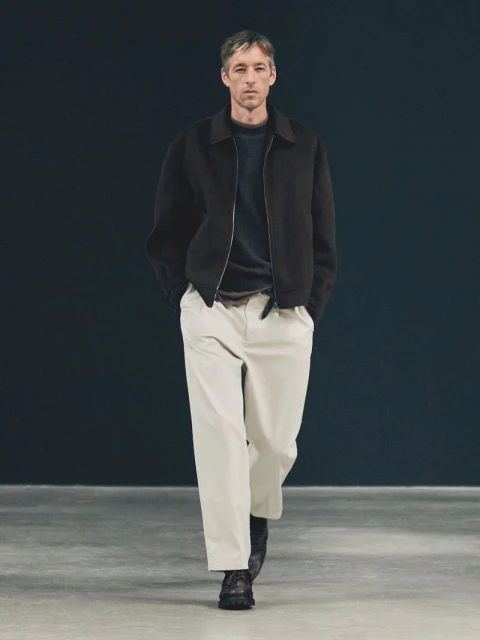 Adult man walks forward in a studio with a dark background wearing a black jacket, sweater, and light-colored pants.