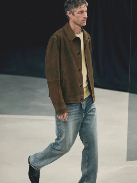 An adult man in a brown suede jacket and jeans walks in a photo studio with a black backdrop and concrete floor, shot in medium perspective.